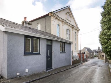 A street with a small gray house with black door and windows next to a large ornate building and other buildings in the background at Ty Capel Bach in Rachub
