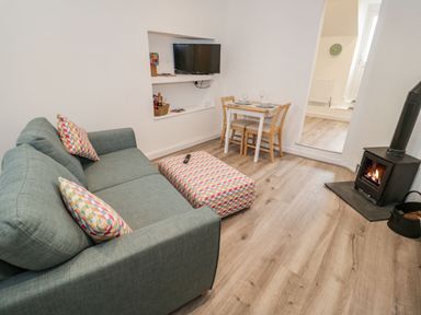 A living room with a green sofa, patterned ottoman, small dining table, wall-mounted TV, and wood burning stove at Ty Capel Bach in Rachub