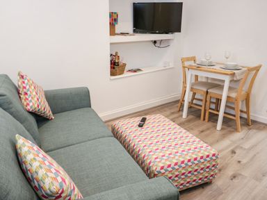 A living room with a green sofa and matching cushions next to a patterned ottoman a wall-mounted TV and a small dining table with two chairs at Ty Capel Bach in Rachub