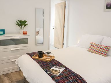 A bedroom with a white bed, a colorful pillow, a dresser with candles and a plant, a wall mirror, and an open door at Ty Capel Bach in Rachub