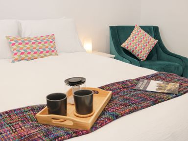 A bedroom with a white bed, colorful pillows, a wooden tray with two black cups, a teapot, a multicolored blanket, a green armchair, and a book at Ty Capel Bach in Rachub