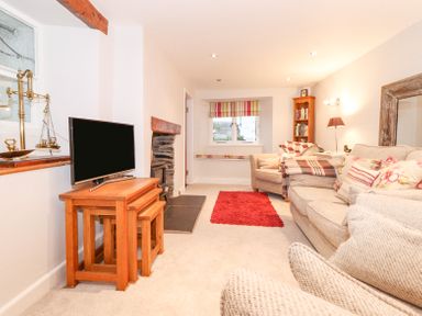 A living room with a television on wooden nesting tables a stone fireplace a red rug armchairs and a sofa with cushions at Yanway Cottage in Troutbeck near Troutbeck Bridge