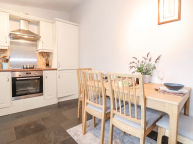 A kitchen with white cabinets and a stainless steel oven next to a wooden dining table with chairs and a plant on the table at Yanway Cottage in Troutbeck near Troutbeck Bridge