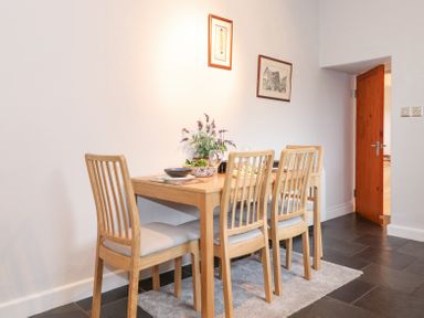 A dining area with a wooden table and four chairs with cushions a plant and dinnerware on the table two pictures on the wall and a wooden door in the background at Yanway Cottage in Troutbeck near Troutbeck Bridge