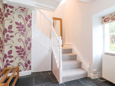 A hallway with stairs carpeted in grey a wooden shoe rack on a tiled floor and floral wallpaper at Yanway Cottage Troutbeck near Troutbeck Bridge