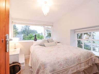 A bedroom with a bed a side table with a lamp and windows showing trees at Yanway Cottage in Troutbeck near Troutbeck Bridge