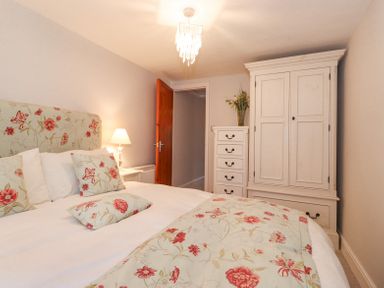 A bedroom with a double bed floral cushions and duvet a white chest of drawers a white wardrobe a wooden door and a bedside lamp at Yanway Cottage in Troutbeck near Troutbeck Bridge