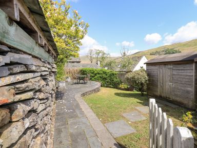 A garden with a stone wall patio area outdoor table and chairs wooden shed and grassy lawn at Yanway Cottage in Troutbeck near Troutbeck Bridge