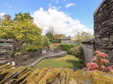 A garden with stone walls a tree bushes a patio with a table and chairs and a wooden shed at Yanway Cottage in Troutbeck near Troutbeck Bridge