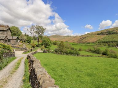 A stone house by a gravel path next to a stone wall with green fields and hills in the background at Yanway Cottage in Troutbeck near Troutbeck Bridge