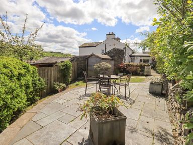 A patio area with a round table and chairs surrounded by plants and a white house in the background at Yanway Cottage in Troutbeck near Troutbeck Bridge