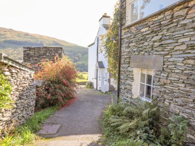 A narrow pathway between stone walls and houses with bushes on the sides and hills in the background at Yanway Cottage in Troutbeck near Troutbeck Bridge