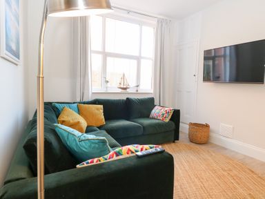 A living room with a green sectional sofa with multicolored cushions a wall-mounted television a floor lamp and a woven basket at Flat 1 in Hunstanton