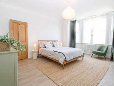 A bedroom with a wooden bed and side tables with lamps a green armchair near windows and a woven rug on the floor at Flat 1 in Hunstanton