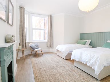 A bedroom with two single beds white bedding and green headboards a chair with cushions a side table a fireplace and large windows with curtains at Flat 1 in Hunstanton