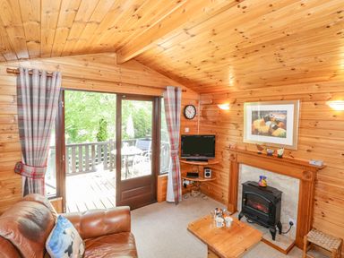 A wooden cabin living room with a leather sofa coffee table fireplace and a TV near a glass door leading to a balcony at Robin's Nest Number 12 Burnside Park near Keswick