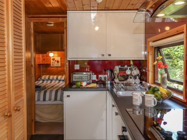 A kitchen with white cabinets a black countertop and a sink next to a window with fruit and two mugs on the counter a small bedroom with a bed and striped bedding at Robins Nest Number 12 Burnside Park near Keswick