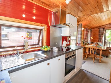 A kitchen with a sink oven stovetop and fruit bowl connected to a dining area with a wooden table chairs and sofa at Robins Nest Number 12 Burnside Park near Keswick