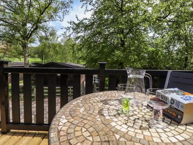A balcony with a mosaic table holding a water pitcher glasses and books overlooking trees and cabins at Robins Nest Number 12 Burnside Park near Keswick