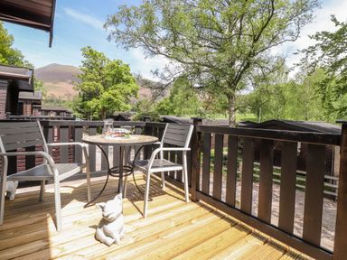 An outdoor balcony with two chairs a round table and a pig statue with trees and hills in the background at Robins Nest in Number 12 Burnside Park near Keswick
