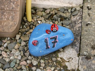 A blue painted rock with number 17 and ladybug decorations on gravel in an outdoor area