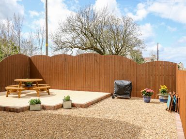 A fenced backyard with gravel ground a round wooden picnic table on a tiled area potted plants and a covered barbecue grill at Stillwaters in Bushmills County Antrim