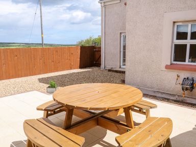 An outdoor patio area with a round wooden picnic table and benches next to a beige house with a window and hose reel at Stillwaters in Bushmills County Antrim