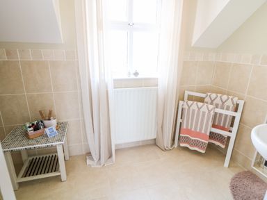 A bathroom corner with a tiled floor and walls a small table with toiletries a towel rack with two towels and a window with light curtains at Stillwaters in Bushmills County Antrim