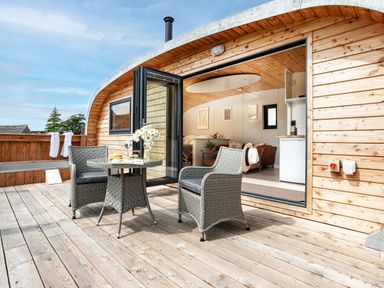 A wooden deck with two wicker chairs and a table outside a small wooden cabin with open doors showing a kitchenette and seating at Low Longlands in Cartmel