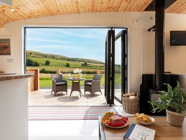 An open living space with a wood stove and a table with snacks looking out to an outdoor patio with two chairs and a table and fields beyond at Low Longlands in Cartmel