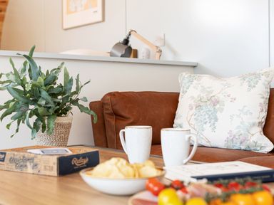 A living room with a brown leather sofa a floral pillow two mugs a potted plant and snacks on a wooden table at Low Longlands in Cartmel