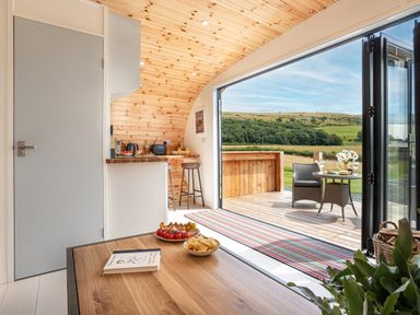 An interior view of a wooden cabin with a kitchen counter and stools next to an open door leading to a wooden deck with chairs and a table overlooking a green field at Low Longlands in Cartmel