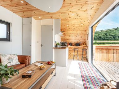A living area with a wooden coffee table, leather sofa, kitchen counter, bar stools, and an open door leading to a wooden deck with green hills outside at Low Longlands in Cartmel