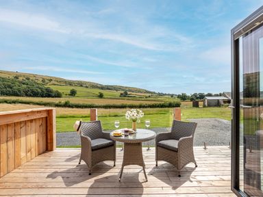 A wooden deck with two wicker chairs and a round table with flowers and wine glasses at Low Longlands in Cartmel