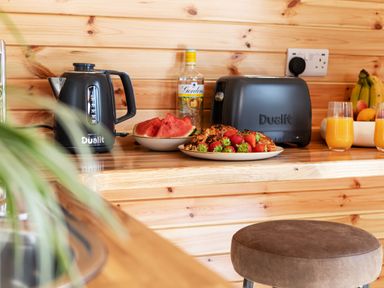 A kitchen countertop with a black kettle, a bottle, plates of watermelon and strawberries with croissants, two glasses of orange juice, and a bowl of fruit at Low Longlands in Cartmel