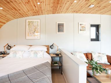 A bedroom with a bed pillows bedside tables lamps wall art a small window and a seating area with cushions and a plant at Low Longlands in Cartmel