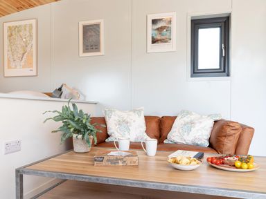 A living area with a brown leather sofa, a wooden table with snacks, two mugs, a plant and framed pictures on the wall at Low Longlands in Cartmel