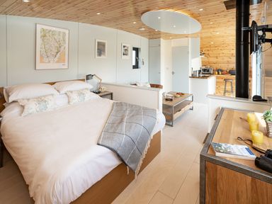 A bedroom with a bed a blanket and pillows a table a small kitchen area a wooden ceiling and light wood flooring at Low Longlands in Cartmel