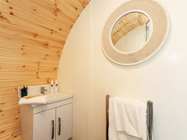 A bathroom corner with a white sink cabinet a round mirror a towel rack with white towels and wooden wall paneling at Low Longlands in Cartmel