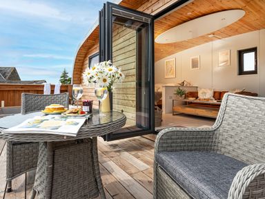 An outdoor wooden deck with a round glass table set with flowers food and drinks next to a room with wooden ceiling and furniture at Low Longlands in Cartmel