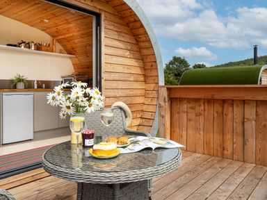 An outdoor wooden deck with a round table set for snacks and drinks next to a wooden pod with a kitchen inside at Low Longlands Cartmel