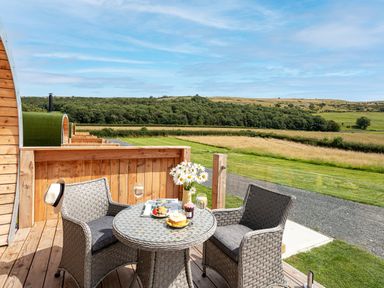A wooden deck with two woven chairs a round table with food and flowers overlooking grass and hills at Low Longlands in Cartmel