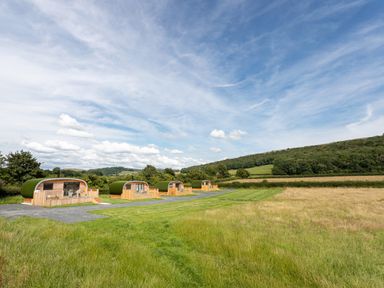 Four wooden cabins with green roofs on a grassy field with hills in the background at Low Longlands in Cartmel