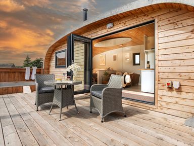 An outdoor wooden deck with two wicker chairs and a round table next to a wooden cabin with an open glass door showing a kitchen and living area at Low Longlands in Cartmel