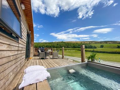 A wooden deck with a hot tub towels table chairs and a barbecue overlooking green fields at Low Longlands in Cartmel