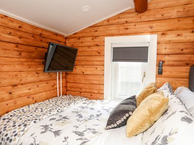 A bedroom with a bed featuring floral bedding and multiple pillows a window with blinds and a mounted TV on wooden walls at Ty Coed - 63 Bronaber near Trawsfynydd