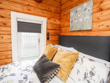 A bedroom with wood paneled walls a window with blinds a bed with pillows and floral bedding and a floral painting above the bed at Ty Coed - 63 Bronaber near Trawsfynydd