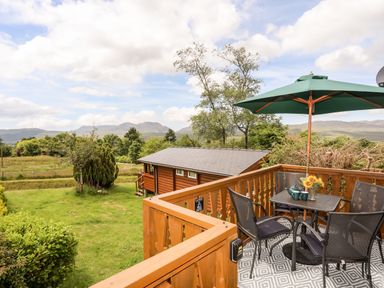 A wooden deck with a table and chairs under a green umbrella overlooking a grassy yard and a wooden cabin at Ty Coed - 63 in Bronaber near Trawsfynydd
