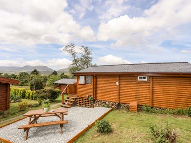An outdoor area with wooden picnic table on gravel next to a wooden cabin in a green landscape at Ty Coed - 63 in Bronaber near Trawsfynydd