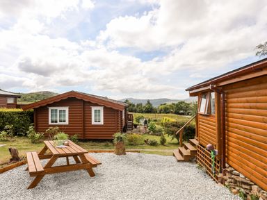 Two wooden cabins with a picnic table on gravel in front and a grassy area with trees and hills in the background at Ty Coed - 63 in Bronaber near Trawsfynydd